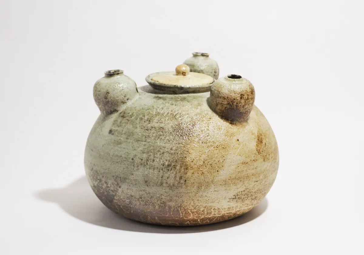 まだら模様とざらついた質感を持つ、蓋付きの壺状作品。全体は灰褐色を帯び、土の荒い表情が残されている。肩部分には3つの小さな球形に近い突起が均等に配置され、それぞれの頂部には丸い穴が開けられている。蓋にも小さなつまみ状の突起があり、全体として素朴さと象徴性を感じさせる造形となっている。作品タイトルの「母」に対応するように、3つの突起が子どもの存在を暗示しているようにも見える。