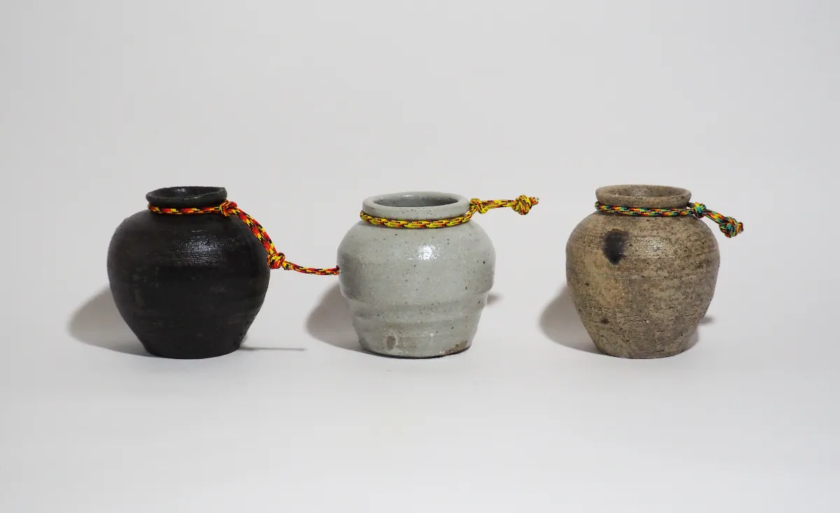 黒、灰白色、灰色の3点で構成された小型の壺作品。壺の首部分には、腰から吊るして持ち歩くことを想起させる紐状の装飾が取り付けられている。作品により、表面には焼成による自然な色むらや質感が見られ、素朴で使い込まれた雰囲気を持つ。実用品と装身具の中間のような、風変わりで遊び心のある印象を与える作品群。