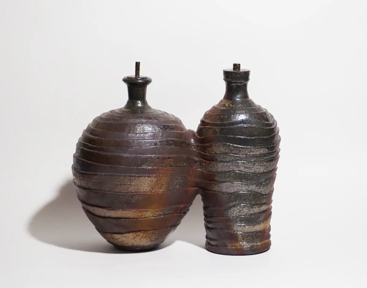 茶褐色と黒褐色の二つの壺が、側面でわずかに融合するように接合された陶芸作品。茶褐色の壺はより丸みを帯びた形状をしており、黒褐色の壺はやや引き締まった印象を与える。両方の壺の表面には、段差を伴う斜め方向の稜線が螺旋状に巡り、器の構造と装飾を同時に形成している。上部には比較的存在感のある首状の造形があり、それぞれの注ぎ口には栓が施され、さらに棒状の装飾が突出している。二つの器が寄り添うように結びつき、対話する人物のような印象を与える作品。