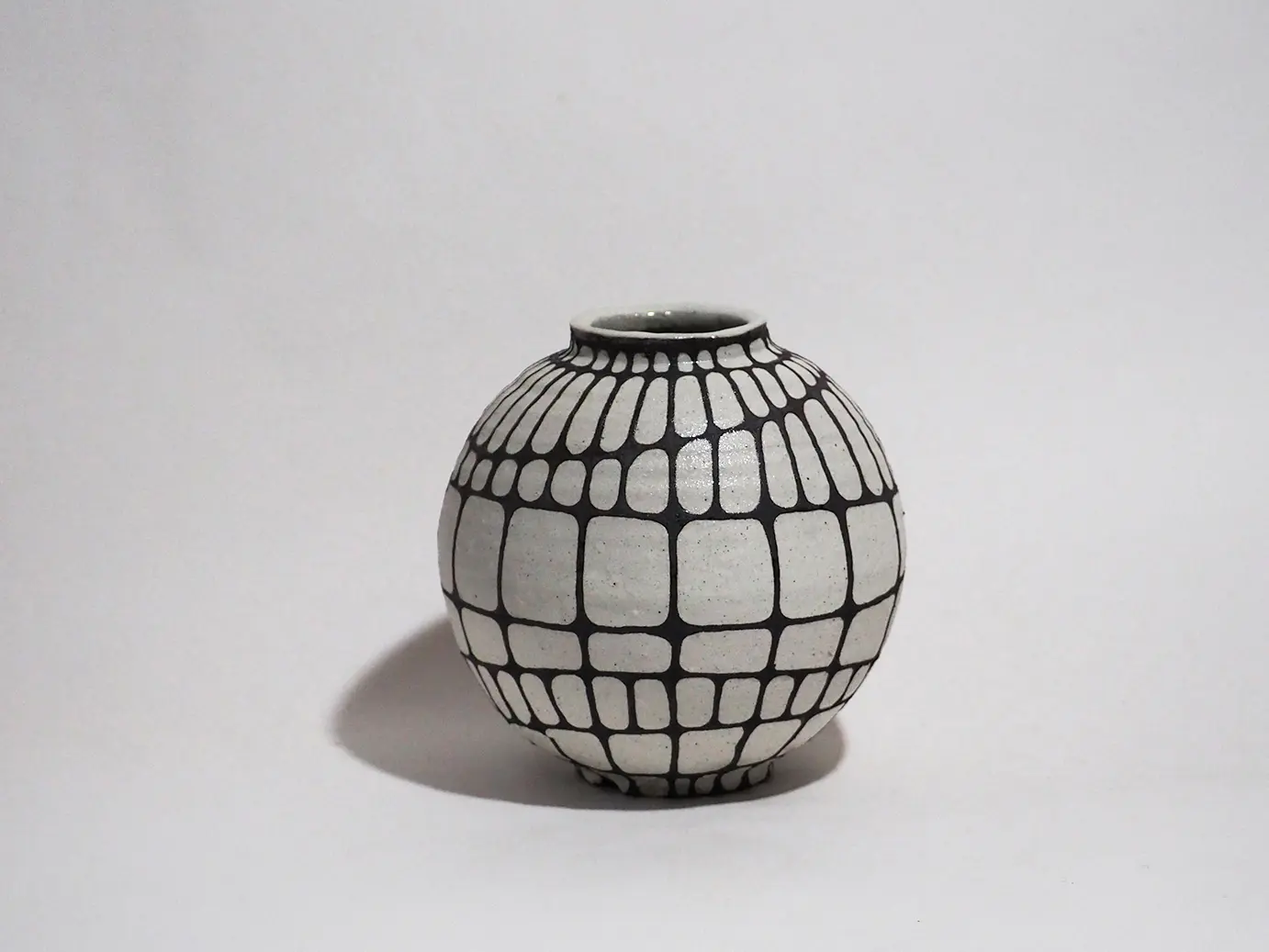灰白色の素地をもつ、球体に近い丸みを帯びた壺状の陶芸作品、表面全体には青色の網目模様が施されており、縦方向と横方向の線が交差している、横方向の線の一部はわずかに斜めに崩れ、柔らかな動きを生んでいる、壺の上部には高さを抑えた短い首状の造形が付けられており、張りのある丸いフォルムと網目模様が強調し合うデザインとなっている。