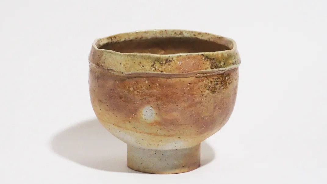 黄褐色を基調とした抹茶茶碗で、表面には自然なまだら模様が現れている。側面は柔らかく丸みを帯びた形状をしており、全体に落ち着いた立ち姿の印象を与える。下部の高台は灰白色で、本体との色の対比が特徴的。口縁付近には折り返し状の造形が施され、手捻りならではの立体感と緊張感を加えている。整った輪郭と、偶然性のある表情が組み合わされた作品。
