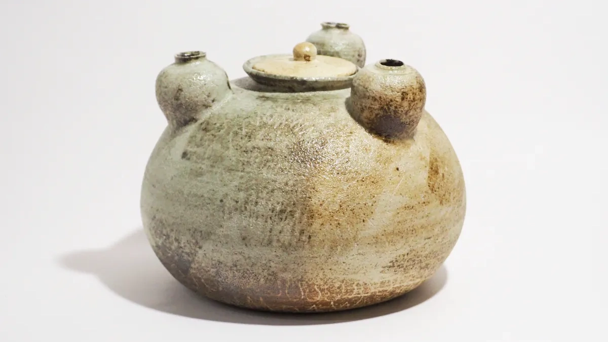 まだら模様とざらついた質感を持つ、蓋付きの壺状作品。全体は灰褐色を帯び、土の荒い表情が残されている。肩部分には3つの小さな球形に近い突起が均等に配置され、それぞれの頂部には丸い穴が開けられている。蓋にも小さなつまみ状の突起があり、全体として素朴さと象徴性を感じさせる造形となっている。作品タイトルの「母」に対応するように、3つの突起が子どもの存在を暗示しているようにも見える。