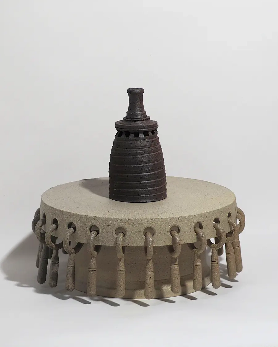 二つの陶芸作品で構成されたセット作品。上部作品は黒褐色の塔状作品で、全体は上に向かって緩やかに細くなる形をしている。側面には螺旋状の溝が巡り、上部には覗き窓を思わせる小さな開口状の造形が施されている。最上部はごく短い円筒形となっており、塔全体に静かな緊張感を与えている。下部作品は灰白色の台座作品で、円盤状の天板を円筒形の支柱が支える構造になっている。天板の側面には多数の穴が開けられており、それぞれに短い鎖やイヤリングのような装飾が吊り下げられている。装飾はわずかに揺れ動く構造となっており、静的な塔作品との対比を生み出している。全体として、重厚な塔と動きのある台座が組み合わされた立体作品となっている。