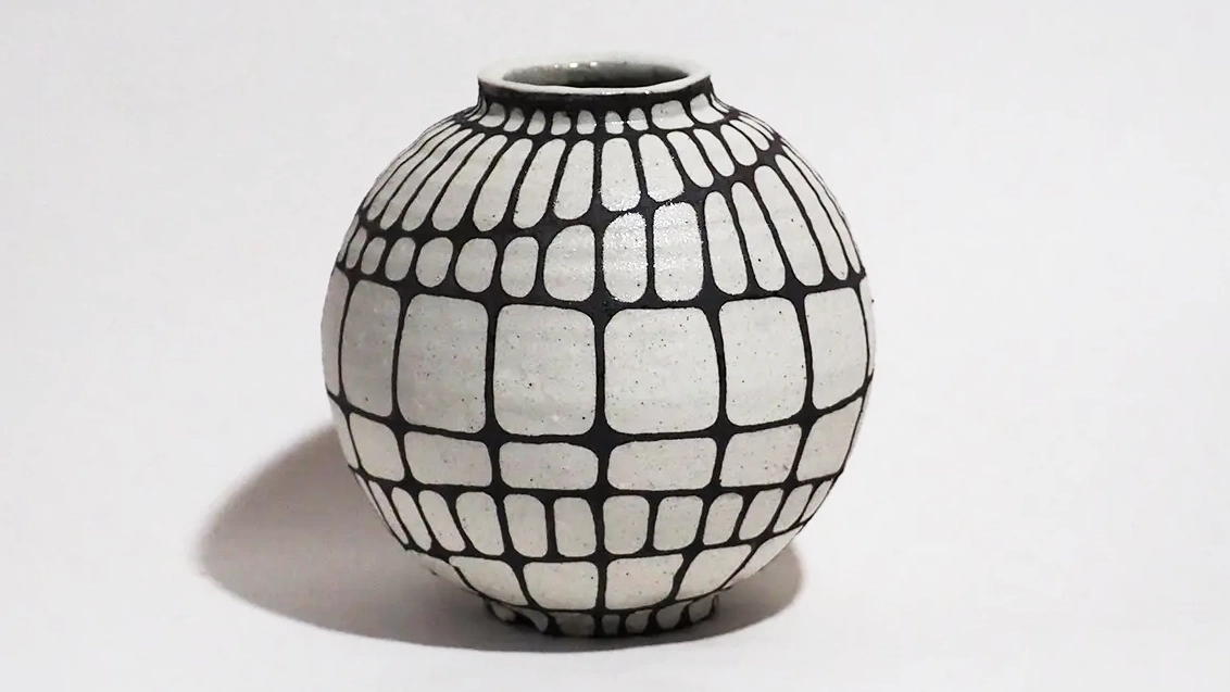 灰白色の素地をもつ、球体に近い丸みを帯びた壺状の陶芸作品、表面全体には青色の網目模様が施されており、縦方向と横方向の線が交差している、横方向の線の一部はわずかに斜めに崩れ、柔らかな動きを生んでいる、壺の上部には高さを抑えた短い首状の造形が付けられており、張りのある丸いフォルムと網目模様が強調し合うデザインとなっている。